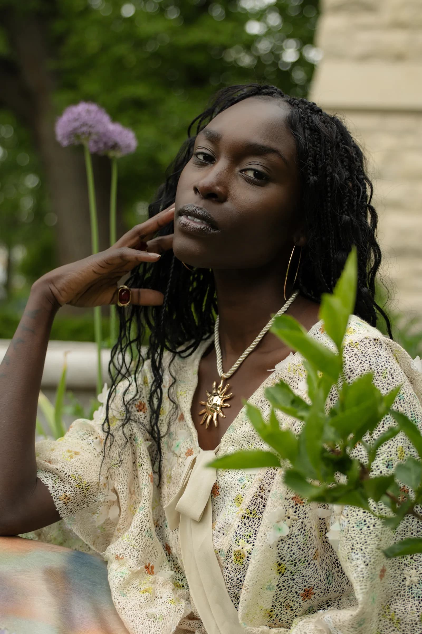A woman in a floral blouse rests her chin on her hand beside tall purple allium.