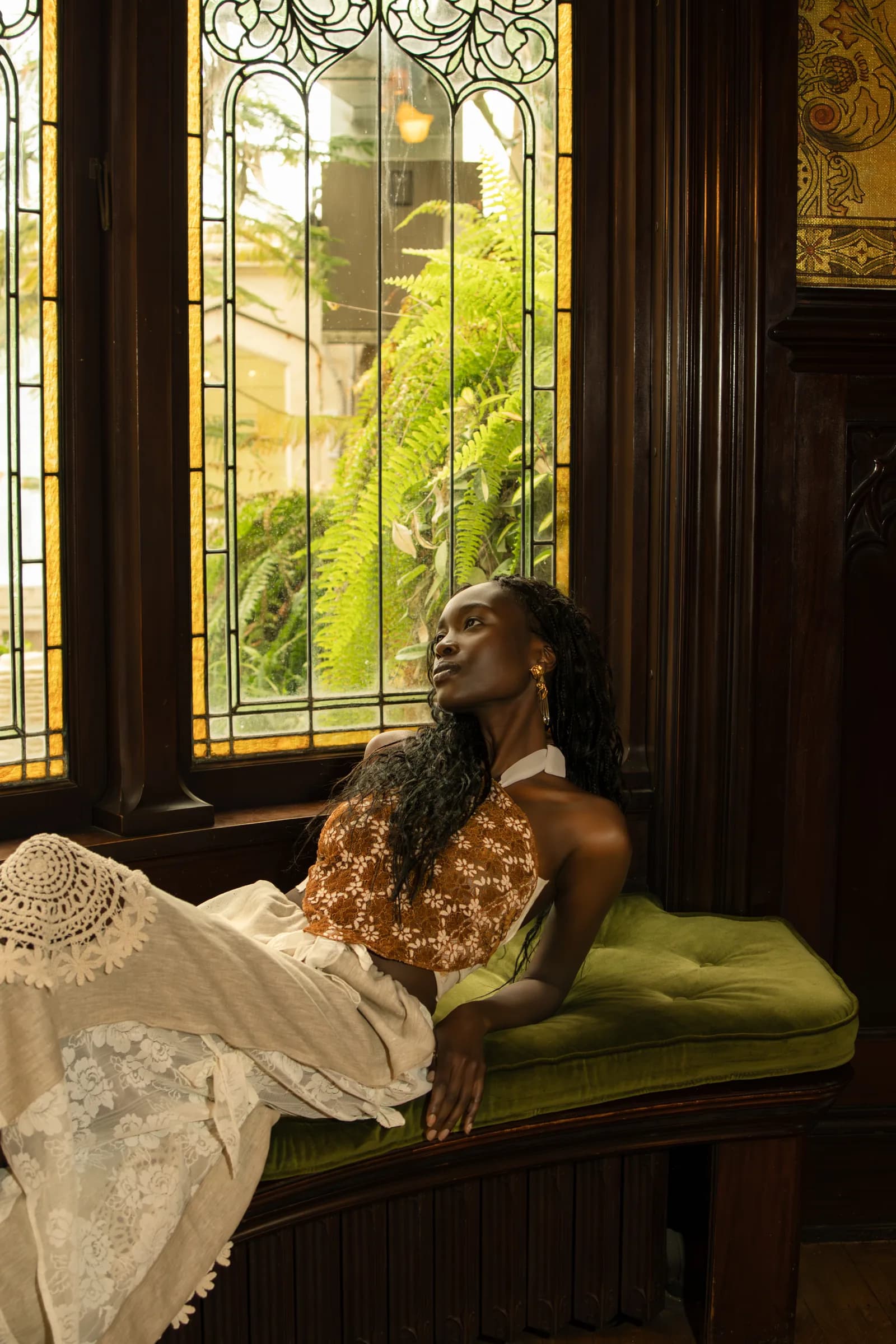 A woman in a white lace dress reclines on a green bench beneath stained glass.