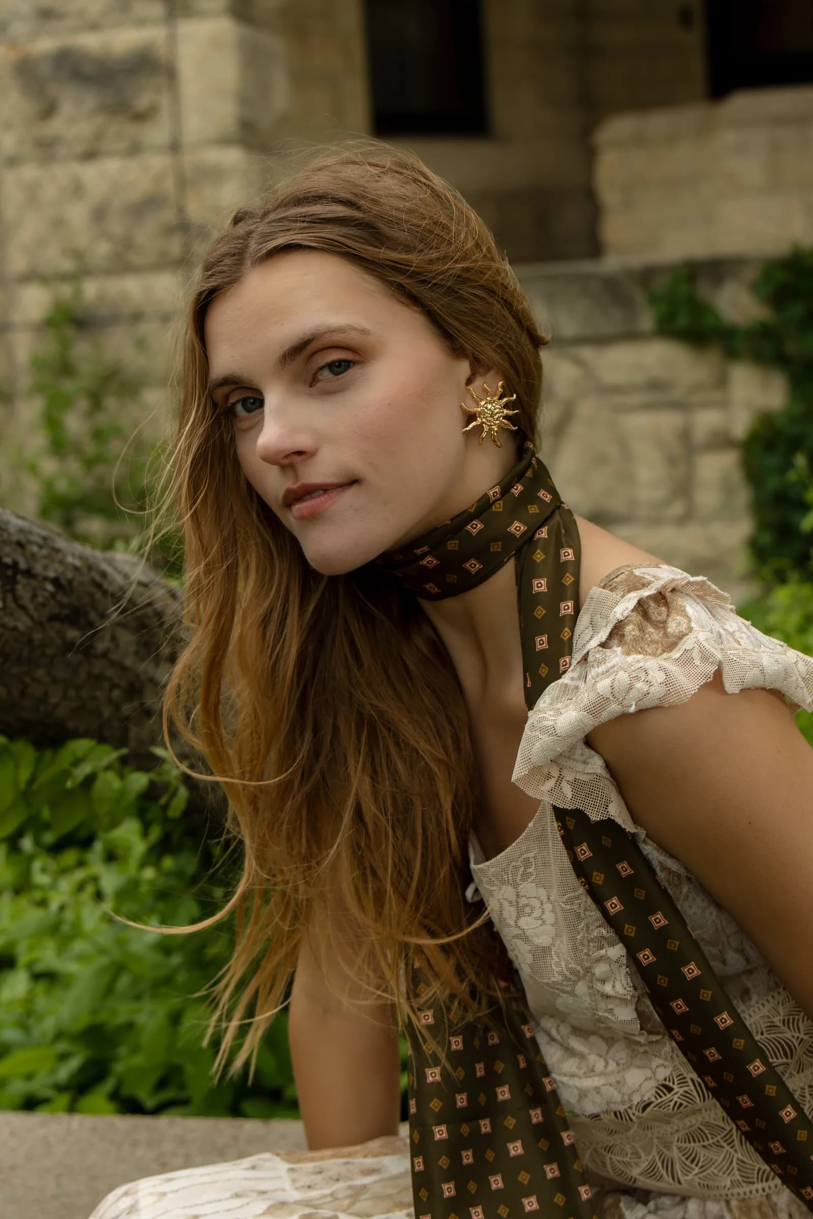 A woman with a long printed scarf looks over her shoulder in front of green leaves.