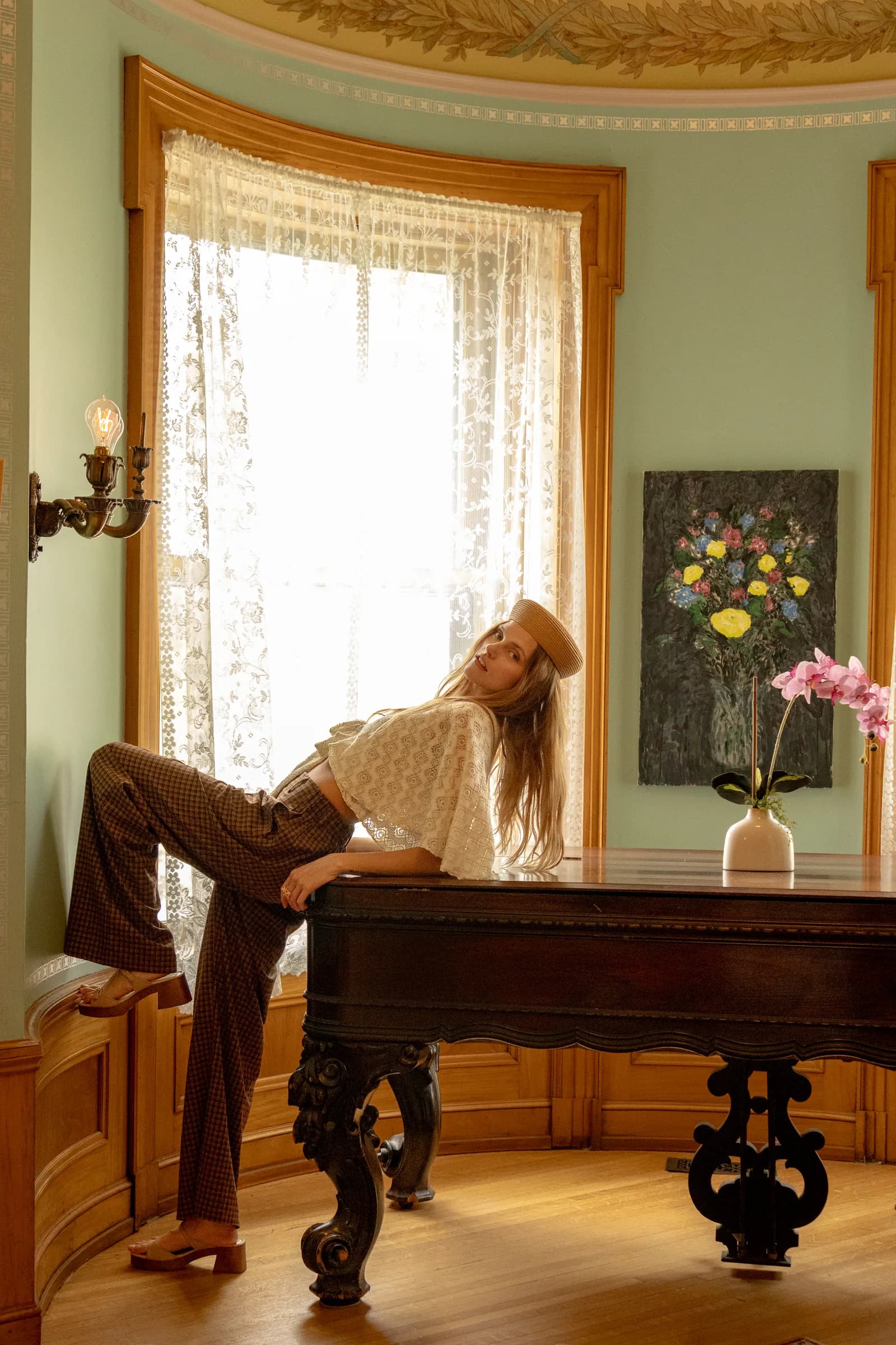 A woman in a cream blouse and dark trousers leans on a carved wooden table by a bright bay window.