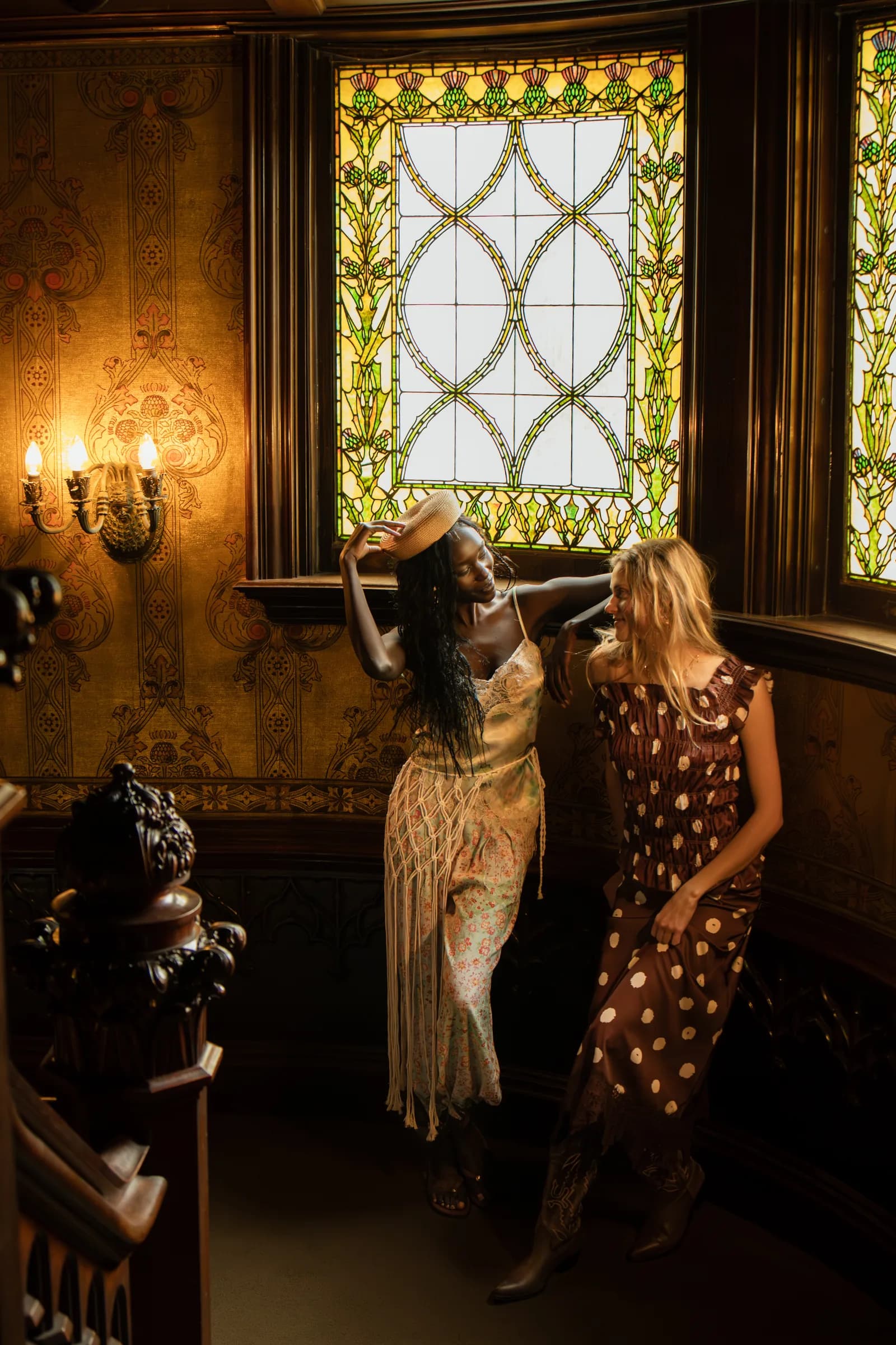 Two women stand together on a landing beneath leaded windows and wall sconces.